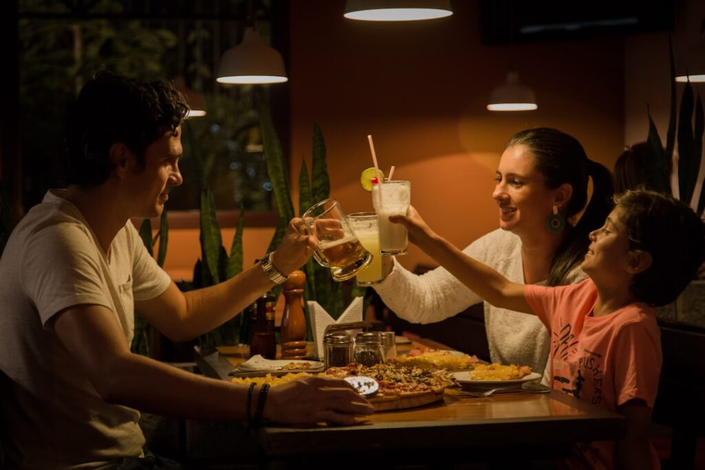 A smiling family shares a meal around a wooden table in a casual restaurant setting with a child holding a plate of food.